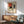 May include: Framed Piet Mondrian exhibition poster with geometric abstract composition in red, blue, yellow, black and white rectangles, displayed above mid-century modern wood credenza with white ceramic lamp, next to sage green upholstered armchair with wooden arms in sunlit room with white walls