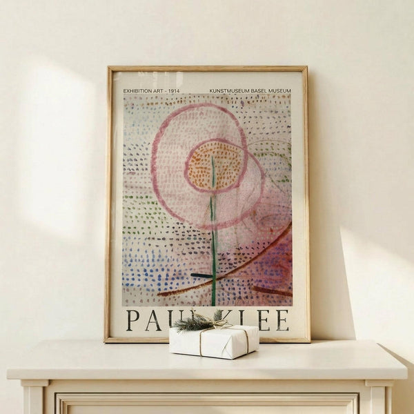 May include: Framed poster with abstract artwork featuring dotted patterns in pink, orange, and blue circles on white background with geometric lines, displayed on light wooden surface next to small white wrapped gift with greenery sprig against cream wall