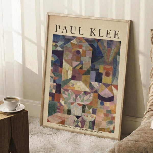 May include: Paul Klee abstract art poster in light wooden frame leaning against white wall. Colorful geometric composition features fragmented shapes in blues, greens, oranges, purples and earth tones with botanical motifs. Museum exhibition poster sits on textured cream rug next to wooden side table with white coffee cup and saucer. Soft natural lighting creates gentle shadows on white paneled walls.