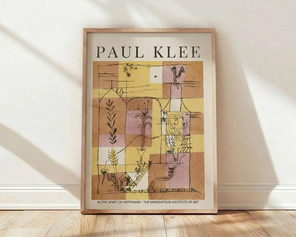 May include: A framed Paul Klee exhibition poster displayed against a white wall on light wood flooring. The artwork features abstract geometric shapes in warm earth tones of yellow, pink, brown, and cream with delicate line drawings of plants, figures, and botanical elements. The poster has "PAUL KLEE" at the top and museum text at the bottom, all within a thin wooden frame. Soft natural lighting creates diagonal shadows across the white textured wall.