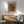 May include: Framed art print of Pierre-Auguste Renoir's floral still life painting featuring pink and red flowers in a blue ceramic pitcher, displayed above a mid-century modern wooden sideboard with white table lamp, next to a sage green upholstered armchair with wooden arms in a sunlit room with white walls