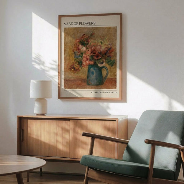 May include: Framed art print of Pierre-Auguste Renoir's floral still life painting featuring pink and red flowers in a blue ceramic pitcher, displayed above a mid-century modern wooden sideboard with white table lamp, next to a sage green upholstered armchair with wooden arms in a sunlit room with white walls