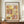 May include: Framed Paul Klee exhibition poster featuring abstract geometric artwork in warm yellows, pinks, and browns with whimsical line drawings of plants and figures. The vintage-style print sits on a wooden sideboard next to a cream ceramic vase and stacked books in a bright room with white paneled walls.