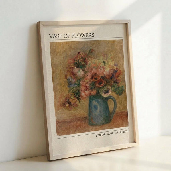 May include: Framed art print featuring Pierre Auguste Renoir's impressionist painting of pink and peach flowers in a blue-green ceramic pitcher. The artwork shows soft, loose brushstrokes with warm golden background tones. White matting surrounds the image with "VASE OF FLOWERS" printed at top and "PIERRE AUGUSTE RENOIR" at bottom. Light wood frame rests against white wall on white surface, casting subtle shadow.
