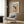 May include: Cream-colored framed art print featuring Jackson Pollock's abstract expressionist drip painting in black, orange, and beige tones, displayed on wood-paneled wall above modern gray upholstered armchair with geometric patterned pillow, brass floor lamp, and dark round side table with open book in contemporary living room setting with natural lighting
