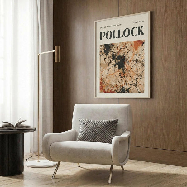 May include: Cream-colored framed art print featuring Jackson Pollock's abstract expressionist drip painting in black, orange, and beige tones, displayed on wood-paneled wall above modern gray upholstered armchair with geometric patterned pillow, brass floor lamp, and dark round side table with open book in contemporary living room setting with natural lighting