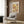 May include: Paul Klee art print in light wood frame hanging on textured brown wood paneling above a cream upholstered armchair with geometric patterned pillow. Abstract artwork features warm yellow, orange and pink rectangular sections with delicate botanical line drawings. Modern brass floor lamp and black side table with open book complete the cozy reading nook scene with natural lighting from nearby window.