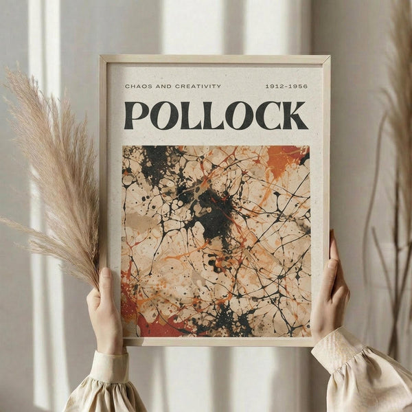 May include: Person in cream ruffled sleeves holding framed abstract art poster featuring Jackson Pollock-style paint splatters and drips in black, orange, and brown tones. Text reads "CHAOS AND CREATIVITY 1912-1956 POLLOCK" above the artwork. Light wood frame against white curtained background with dried pampas grass visible on left side.