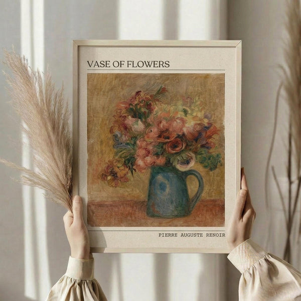 May include: Person holding framed art print showing impressionist-style floral still life painting with pink and coral flowers in blue-green ceramic pitcher against golden brown background. Natural light streams through white curtains, dried pampas grass visible on left. Warm, neutral tones throughout scene.
