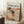 May include: Framed art print leaning against white wall corner featuring Jackson Pollock's signature drip painting style with chaotic black lines, orange and red paint splatters on cream background. Print reads "CHAOS AND CREATIVITY" and "POLLOCK 1912-1956" at top. Natural wood frame, positioned on textured white rug next to wooden side table with white coffee cup, beige throw blanket visible at right edge.