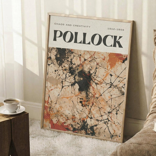 May include: Framed art print leaning against white wall corner featuring Jackson Pollock's signature drip painting style with chaotic black lines, orange and red paint splatters on cream background. Print reads "CHAOS AND CREATIVITY" and "POLLOCK 1912-1956" at top. Natural wood frame, positioned on textured white rug next to wooden side table with white coffee cup, beige throw blanket visible at right edge.