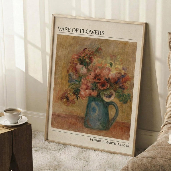 May include: Framed art print leaning against white wall corner on fluffy white rug, showing colorful floral painting of roses and flowers in blue ceramic pitcher. Warm impressionist style with pink, orange, and green blooms against golden background. White coffee cup on wooden block nearby, beige throw blanket visible at edge.