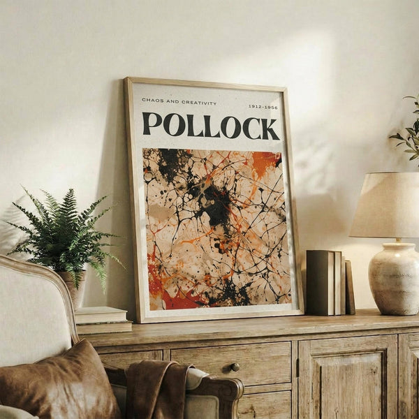 May include: Framed Jackson Pollock exhibition poster featuring abstract drip painting with black paint splatters, orange and beige tones, and chaotic linear marks on cream background. Light wood frame sits on rustic wooden sideboard next to fern plant, books, and ceramic vases in warm neutral living room setting.