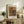 May include: Framed Renoir floral artwork showing pink and red flowers in blue ceramic vase, displayed on rustic wooden sideboard alongside fern plant, books, ceramic jar and table lamp in warm-toned living room setting with beige upholstered chair visible in foreground.