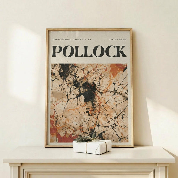May include: Framed art poster featuring Jackson Pollock's abstract expressionist drip painting with black, orange, and beige paint splatters and lines on cream background. Text reads "CHAOS AND CREATIVITY" and "POLLOCK" with dates "1912-1956." Light wood frame sits on white wooden surface next to small wrapped gift with twine and evergreen sprig against cream wall.