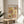 May include: Framed Paul Klee exhibition poster featuring abstract geometric artwork in warm yellow, pink, and brown tones with botanical line drawings, displayed on light wood round side table next to beige upholstered chair and plant leaves in sunlit corner with white walls