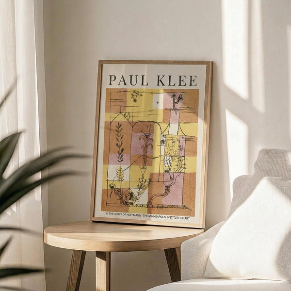 May include: Framed Paul Klee exhibition poster featuring abstract geometric artwork in warm yellow, pink, and brown tones with botanical line drawings, displayed on light wood round side table next to beige upholstered chair and plant leaves in sunlit corner with white walls