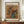 May include: Framed reproduction of Renoir's floral still life painting showing colorful flowers in a blue ceramic pitcher, displayed on a wooden sideboard next to a small white vase and books. The artwork features warm tones with pink, red, and yellow blooms against a golden brown background, presented in a thin brown frame with white matting and black text identifying the title and artist.