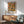 May include: Abstract expressionist art print with vibrant orange, blue, yellow and green paint splatters and black lines in wooden frame hanging above mid-century modern wooden credenza with white ceramic lamp, next to sage green upholstered armchair with wooden arms in sunlit room with white walls