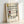 May include: Framed art print featuring Piet Mondrian's geometric abstract composition with black grid lines and rectangular blocks in yellow, red, and blue on cream background. Artist name and dates "1872-1944" with "Trafalgar Square" printed below artwork. Light wood frame leaning against white wall, showing natural lighting and shadows on the glass surface.