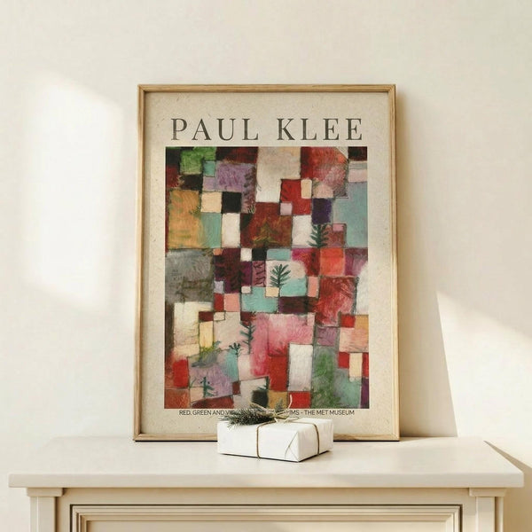 May include: Framed poster featuring Paul Klee's abstract geometric artwork with colorful squares and rectangles in reds, greens, blues, and earth tones, displayed on a light wood shelf against a cream wall. Small white gift box with twine and evergreen sprig sits in front of the natural wood frame.