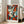 May include: Framed abstract cubist-style portrait featuring fragmented face with geometric shapes in vibrant orange, red, teal, and cream colors. The artwork shows angular facial features with prominent eye and lips, displayed in light wood frame leaning against white wall corner next to beige upholstered chair in bright, naturally-lit room with large windows.