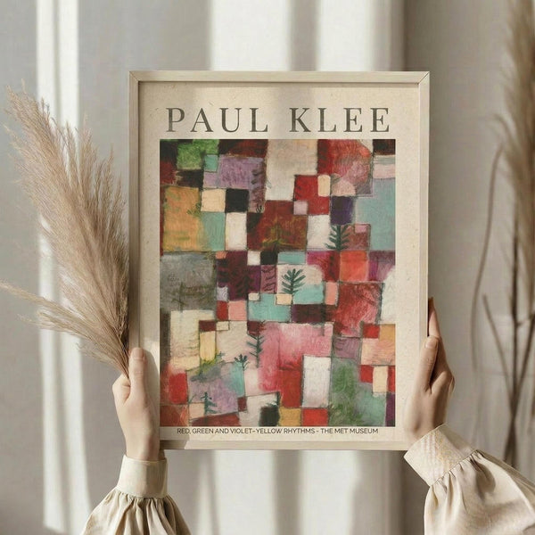 May include: Person holding framed Paul Klee art print featuring colorful geometric squares in reds, greens, purples, and yellows with small tree motifs. Light wood frame against neutral beige clothing and dried wheat stems. Soft natural lighting creates warm, minimalist styling.