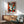 May include: Framed geometric abstract portrait print in cubist style hanging above wood credenza. Artwork features stylized face with blue eye rendered in angular orange, teal, and red segments. Mid-century modern room setting with sage green upholstered chair, white ceramic table lamp, and natural wood furniture. Sunlight casts dramatic shadows across white wall.