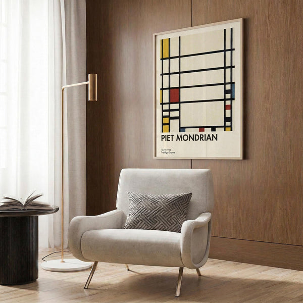 May include: Framed Piet Mondrian abstract art print featuring geometric black lines with yellow and red rectangles on cream background, displayed on wood paneled wall above beige upholstered chair with patterned pillow, brass floor lamp and side table with open book nearby