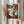 May include: Hands holding a framed abstract portrait print in cubist style, featuring geometric shapes in red, orange, blue, and green tones forming a stylized face with prominent eye and lips. Light wood frame against neutral curtained background with dried pampas grass visible on left side.