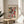 May include: Framed Paul Klee abstract art print displayed on round wooden side table next to beige upholstered chair. The colorful geometric artwork features mosaic-like squares in red, green, violet, yellow, and other vibrant hues with small tree motifs scattered throughout. Natural sunlight streams across the neutral-toned room setting with plant leaves visible in corner.