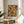 May include: Abstract expressionist poster with vibrant orange, blue, yellow and green paint splatters and black lines in wooden frame, displayed on round wooden side table next to beige upholstered chair. Sunlight streams across white wall creating soft shadows. Green plant leaves visible in corner of minimalist modern living room setting.