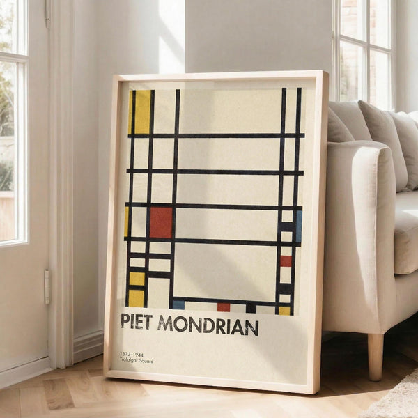 May include: Framed art print featuring geometric abstract design with black grid lines and colored rectangles in yellow, red, and blue on cream background. Shows artist name "PIET MONDRIAN" and dates "1872-1944 Trafalgar Square" below artwork. Light wood frame leaning against white wall next to beige upholstered sofa in bright, naturally lit room with hardwood floors.
