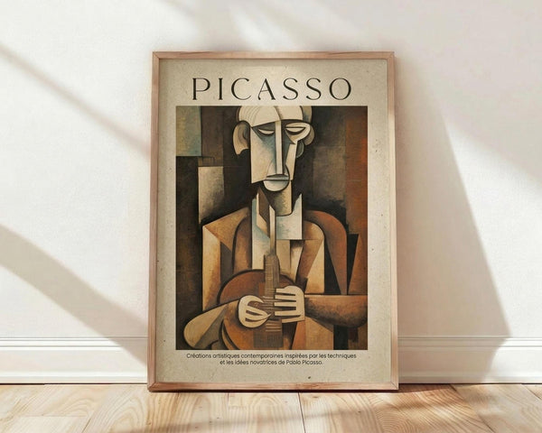 May include: Framed Picasso cubist artwork poster showing abstract musician figure in earth tones - browns, beiges, and muted colors. The geometric, fragmented portrait depicts a seated figure holding what appears to be a musical instrument. Poster has cream border with "PICASSO" text at top and French description at bottom. Displayed in light wooden frame leaning against white wall on natural wood floor with soft window lighting casting shadows.