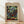 May include: Framed art print featuring Paul Klee's abstract mosaic composition of colorful geometric squares in greens, blues, reds, yellows, and earth tones arranged in a grid pattern. The poster has a cream-colored border with "PAUL KLEE" text at top and "MAY PICTURE - THE MET MUSEUM" at bottom. Natural wood frame leaning against white wall on light wooden floor with soft window light casting shadows.