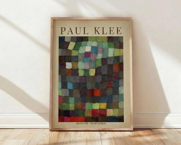 May include: Framed art print featuring Paul Klee's abstract mosaic composition of colorful geometric squares in greens, blues, reds, yellows, and earth tones arranged in a grid pattern. The poster has a cream-colored border with "PAUL KLEE" text at top and "MAY PICTURE - THE MET MUSEUM" at bottom. Natural wood frame leaning against white wall on light wooden floor with soft window light casting shadows.
