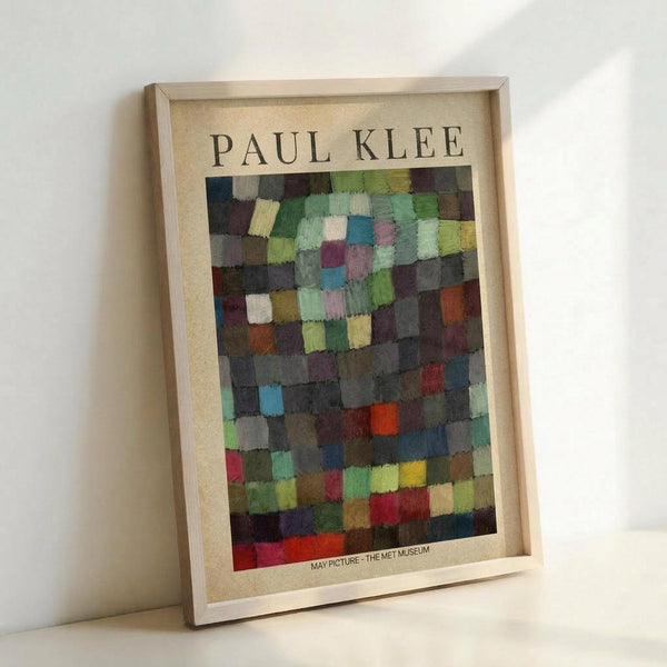 May include: Framed poster of Paul Klee's abstract artwork featuring a mosaic-like grid of colorful painted squares in greens, blues, purples, reds, and earth tones. The print has a cream-colored border with "PAUL KLEE" at the top and "MAY PICTURE - THE MET MUSEUM" at the bottom. Displayed in a light wooden frame leaning against a white wall on a light surface.