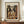 May include: Framed Picasso-style cubist poster showing abstract musician figure in brown, beige and orange geometric shapes holding stringed instrument. French text below artwork. Displayed on wooden console table with white ceramic vase and books in elegant room with cream walls and white molding.