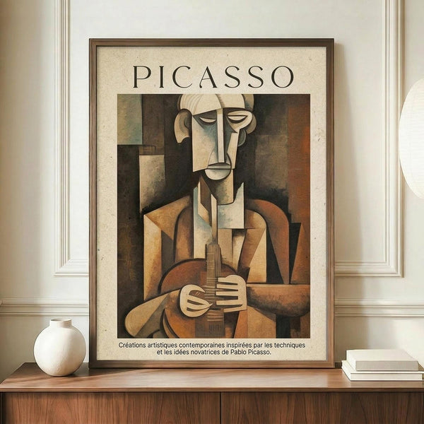 May include: Framed Picasso-style cubist poster showing abstract musician figure in brown, beige and orange geometric shapes holding stringed instrument. French text below artwork. Displayed on wooden console table with white ceramic vase and books in elegant room with cream walls and white molding.