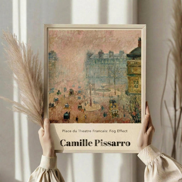 May include: Framed art print of Camille Pissarro's impressionist street scene showing a misty Parisian square with carriages, pedestrians, and Haussmann-style buildings in muted pinks and blues. Person in cream sweater holds the beige-framed poster while standing near dried pampas grass against white curtained window, creating soft natural lighting.