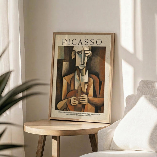 May include: Framed Picasso cubist artwork print displayed on round wooden side table next to beige upholstered chair. The poster shows an angular, geometric figure of a seated musician in earth tones of brown, orange, and cream. Natural sunlight streams through window creating soft shadows. Green plant leaves visible in corner. Modern minimalist home decor setting with neutral color palette.