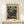 May include: Framed Paul Klee abstract art print featuring a mosaic of colorful squares in muted greens, blues, purples, reds and earth tones arranged in a grid pattern. The poster has a cream colored mat and thin wooden frame, displayed on a light wood surface alongside a small white wrapped gift with natural twine and greenery against a white wall.