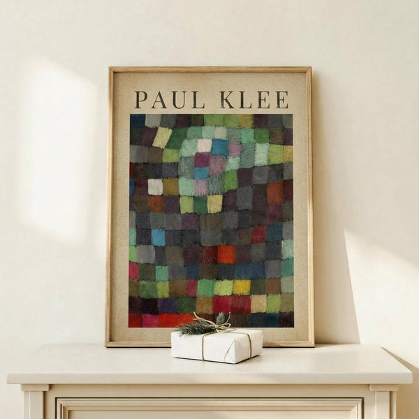 May include: Framed Paul Klee abstract art print featuring a mosaic of colorful squares in muted greens, blues, purples, reds and earth tones arranged in a grid pattern. The poster has a cream colored mat and thin wooden frame, displayed on a light wood surface alongside a small white wrapped gift with natural twine and greenery against a white wall.