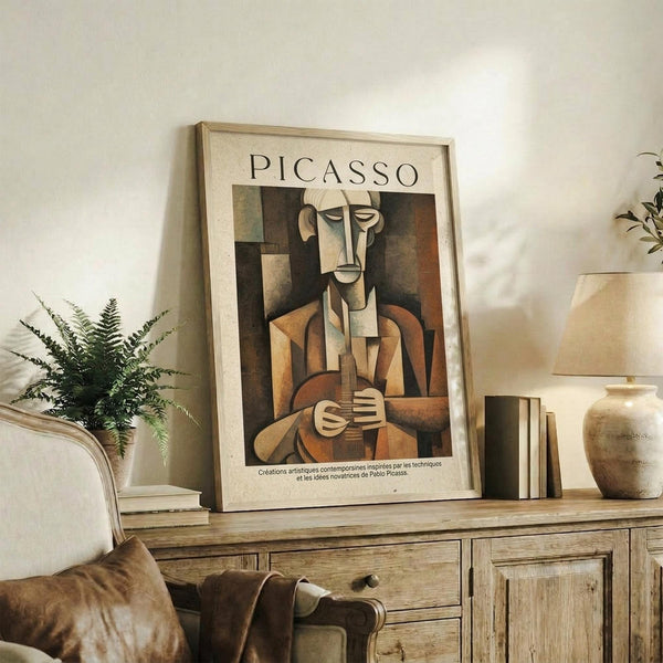 May include: Framed Picasso cubist artwork showing abstract musician figure in earth tones of brown, orange and beige, displayed on rustic wooden console table alongside fern plant, ceramic vase, and books in neutral-toned living room setting with cream upholstered chair