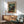 May include: Colorful abstract grid artwork by Paul Klee displayed in wooden frame above mid-century modern credenza with table lamp. Sage green upholstered armchair with wooden frame sits nearby in bright, naturally lit room with white walls and warm wood furnishings.