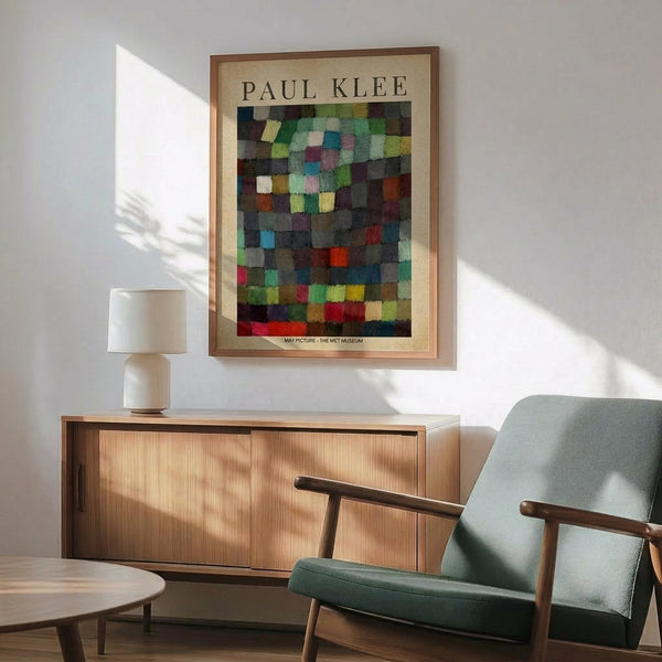 May include: Colorful abstract grid artwork by Paul Klee displayed in wooden frame above mid-century modern credenza with table lamp. Sage green upholstered armchair with wooden frame sits nearby in bright, naturally lit room with white walls and warm wood furnishings.