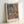 May include: Framed art print in light wood frame leaning against white wall, showing colorful portrait of woman in traditional dress with red and blue striped outfit, ornate headpiece, and folk costume details painted in cubist-inspired style with muted blues, oranges, and earth tones in background