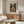 May include: Picasso cubist artwork poster displayed above a wooden mid-century modern credenza in a minimalist living room. The beige and brown toned print shows an angular, geometric figure holding an instrument. White sculptural chair and glass oval coffee table visible in foreground with natural lighting creating shadows across the scene.