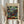 May include: Person in beige clothing holding framed Paul Klee abstract art print featuring colorful mosaic-like squares in greens, blues, purples, reds and yellows. Light wood frame with museum attribution text below artwork. Dried wheat stems visible on left side against soft neutral curtain background.
