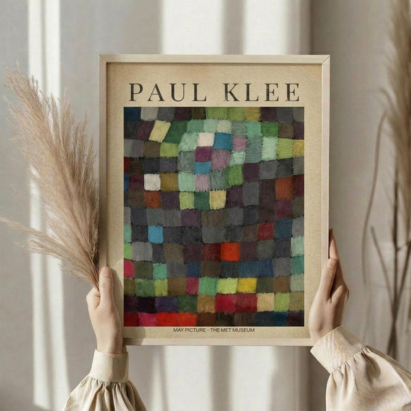 May include: Person in beige clothing holding framed Paul Klee abstract art print featuring colorful mosaic-like squares in greens, blues, purples, reds and yellows. Light wood frame with museum attribution text below artwork. Dried wheat stems visible on left side against soft neutral curtain background.